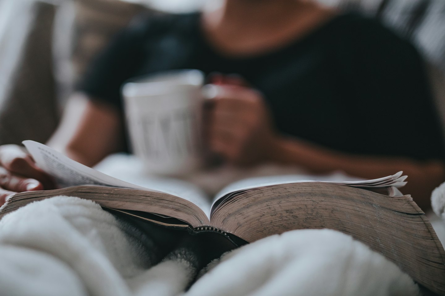 Hands holding a book and a mug on a blanket.